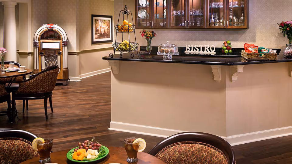 Interior view of a dining area in a senior living facility featuring a wooden floor, a counter with a black countertop labeled 'BISTRO', a tiered fruit stand, a glass cake stand with pastries, baskets with chips, and a table with two glasses of iced tea garnished with lemon slices and a plate of cheese and grapes. In the background, there is a vintage jukebox, framed artwork on the wall, and upholstered chairs around tables.