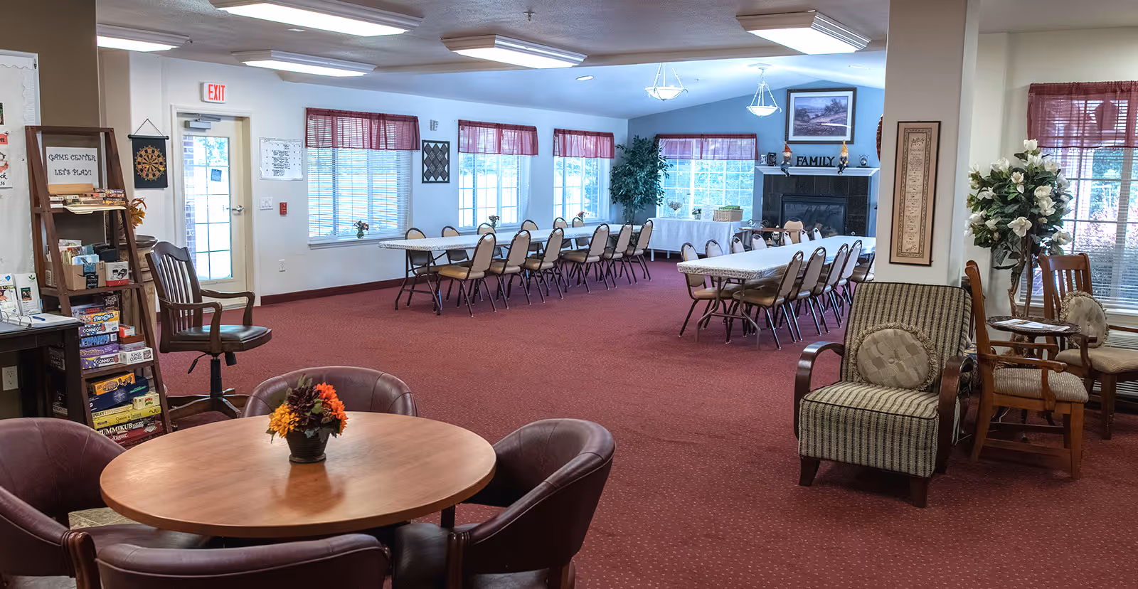 A spacious common area in a senior living facility with red carpet flooring and multiple seating arrangements including round tables with chairs and long tables with chairs. The room has large windows with red valances, a fireplace with a 'FAMILY' sign above it, and a game center shelf with board games and a dartboard on the wall. There are also decorative plants and framed artwork on the walls.