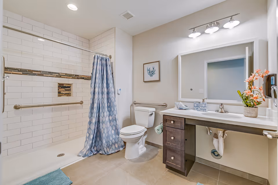 A clean and modern bathroom with a walk-in shower featuring white subway tiles and a decorative tile strip. The shower has a blue and white patterned curtain and a grab bar. There is a white toilet next to the shower, and a vanity with a sink, dark wood cabinets, a large mirror, and a light fixture with three bulbs above it. A small framed artwork and a potted plant are on the wall and countertop respectively.