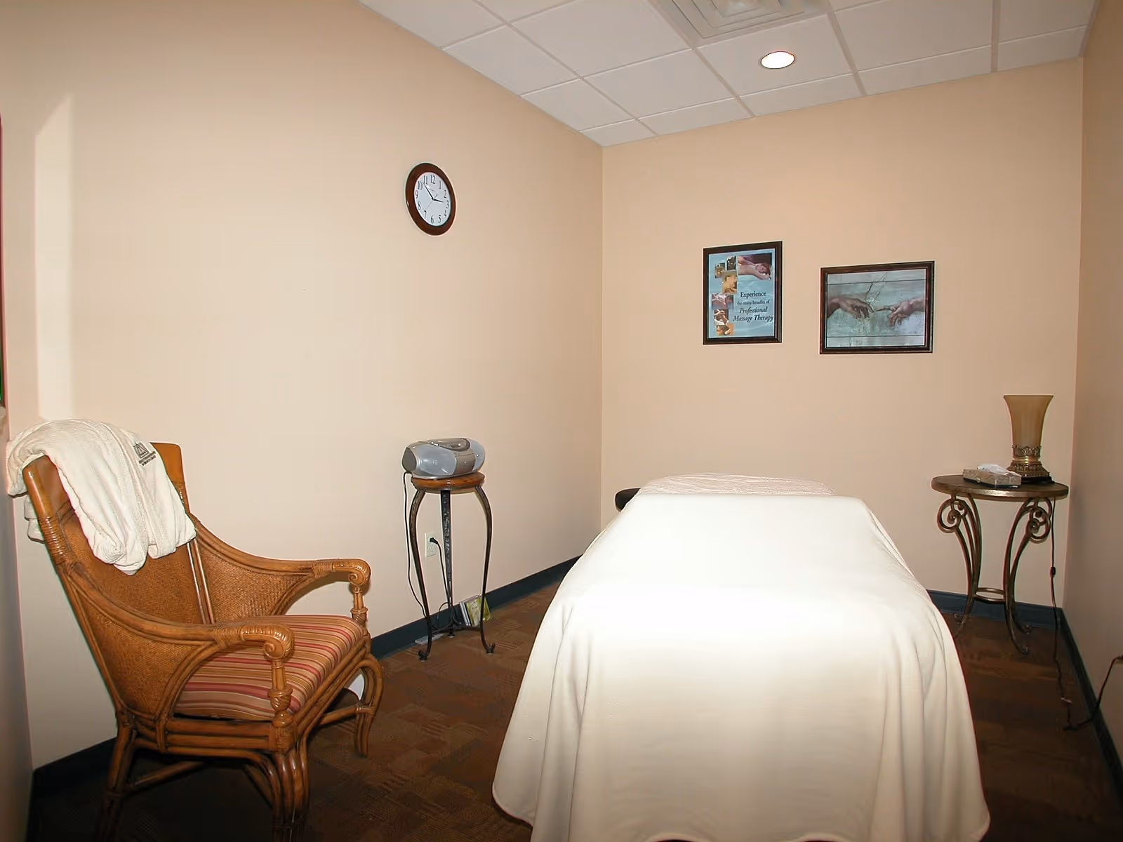 A small room with beige walls featuring a massage table covered with a white sheet in the center. To the left is a wooden chair with a striped cushion and a white towel draped over the back. On the right side, there is a small round table with a decorative lamp and a box of tissues. Two framed pictures hang on the wall above the massage table, and a clock is mounted on the wall above the chair. A small stereo is placed on a tall stand near the corner.