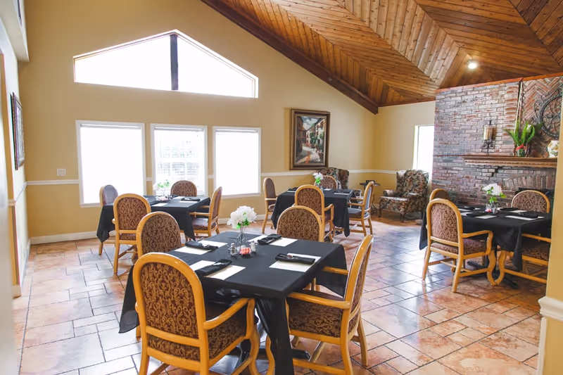 A dining room with several tables covered in black tablecloths, each set with napkins and utensils. The room has large windows letting in natural light, a high wooden ceiling, tiled floor, and a brick fireplace with decorative items. There are upholstered chairs around the tables and a couple of armchairs near the fireplace.