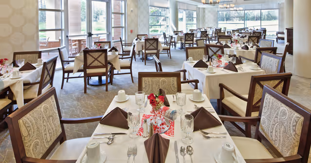 A bright dining room with multiple tables set for a meal. Each table is covered with a white tablecloth and has brown folded napkins, glassware, cups, and silverware neatly arranged. The chairs have wooden frames with patterned upholstery. Large windows allow natural light to fill the room, and there are decorative centerpieces on the tables.