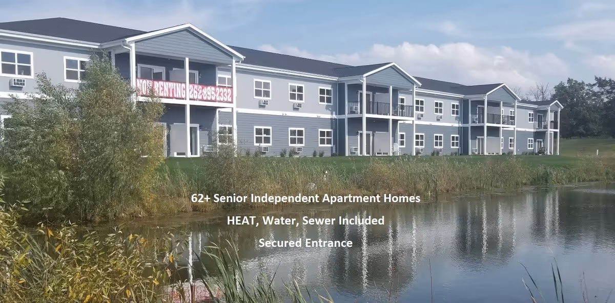A two-story senior independent apartment building with blue siding and white trim, situated behind a pond with reflections of the building in the water. There is greenery including trees and bushes around the pond. A banner on the building reads 'NOW RENTING 262-895-2332'. Text overlay on the image states '62+ Senior Independent Apartment Homes, HEAT, Water, Sewer Included, Secured Entrance'.