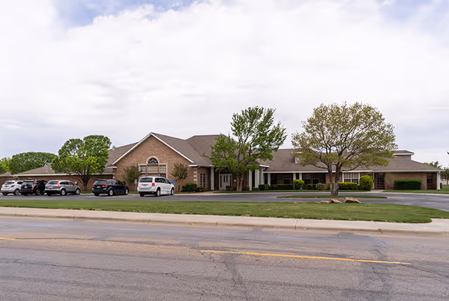 Exterior view of a single-story brick building with a pitched roof, several trees, and parked cars in front, under a cloudy sky.