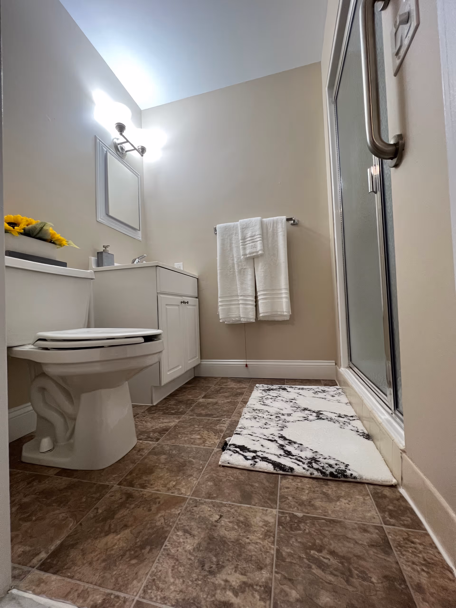 A clean bathroom with a white toilet, a white vanity with a sink, a mirror above the sink, a wall-mounted light fixture, a towel rack with white towels, a glass shower door, and a white and black patterned bath mat on a brown tiled floor.