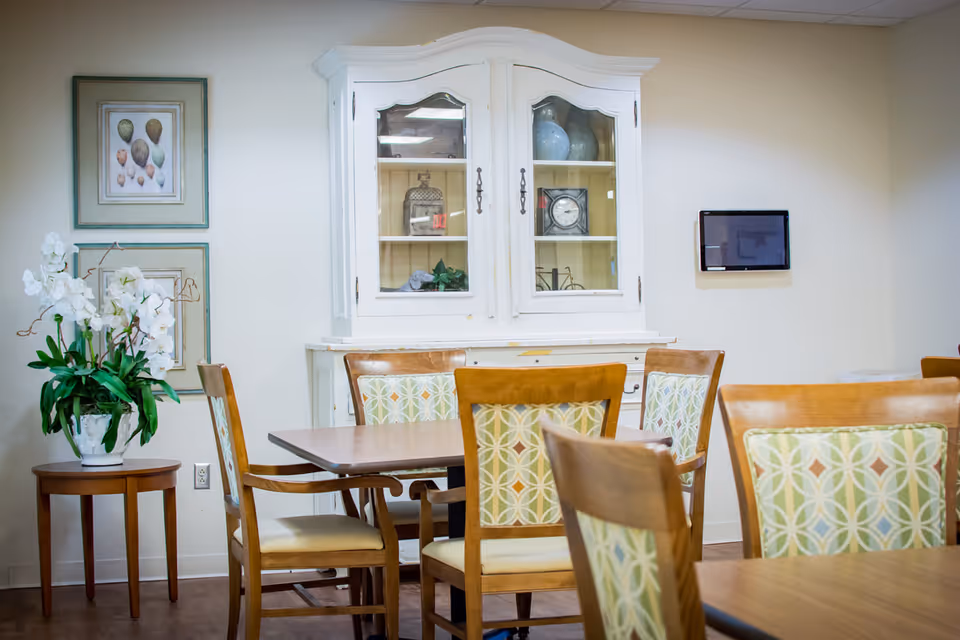 A bright dining area with wooden tables and patterned chairs, a white hutch, wall art, and a potted orchid.