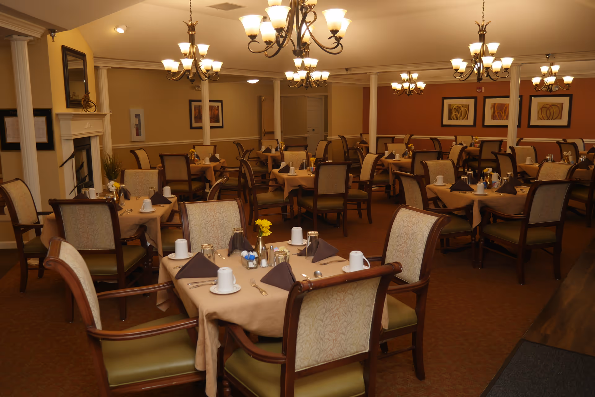 A warmly lit dining room with multiple tables set with cups, napkins, and silverware beneath chandeliers.