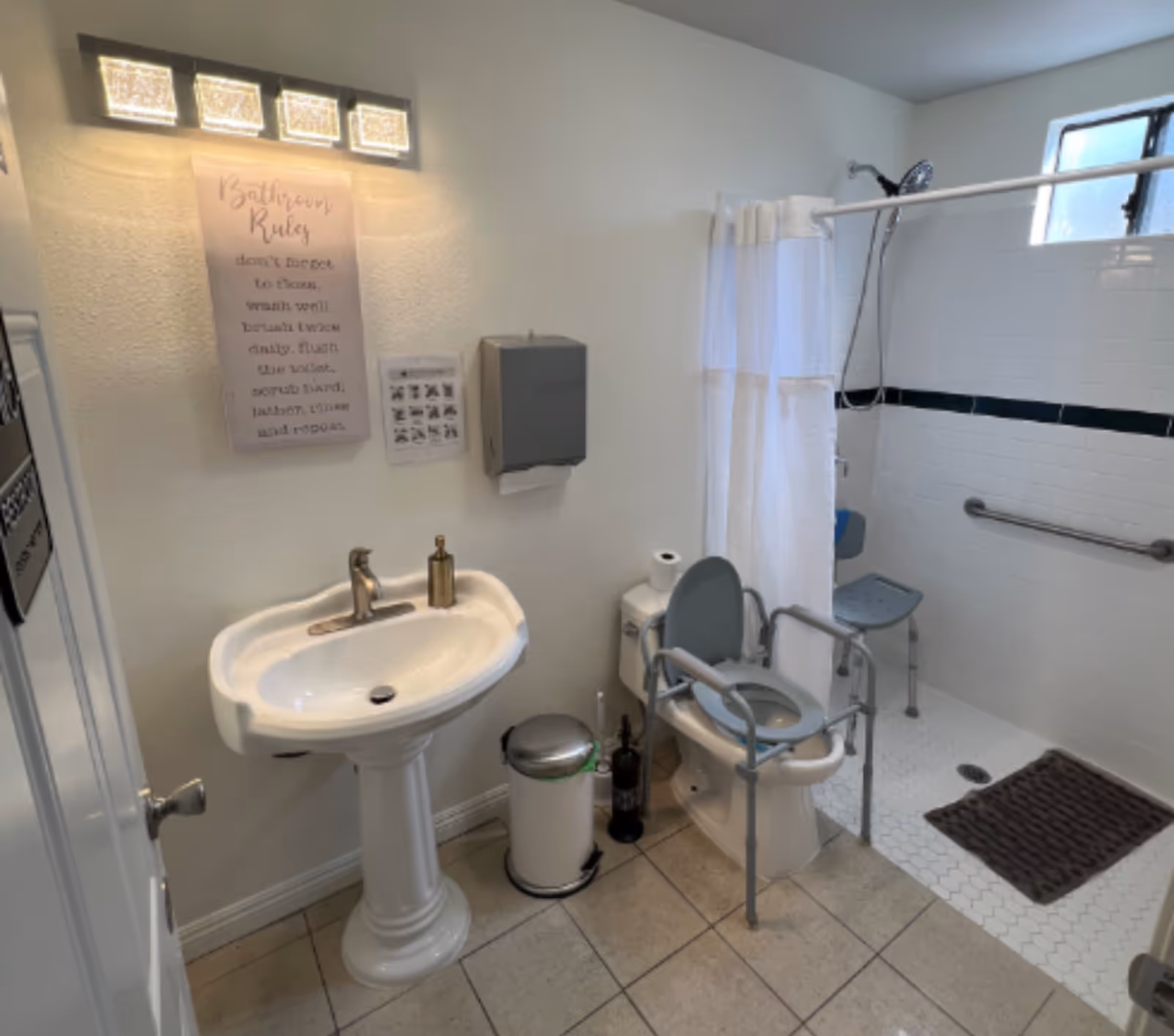 A clean, accessible bathroom with a pedestal sink, a toilet equipped with safety rails and a raised seat, a walk-in shower with a shower chair and grab bars, a small window, a trash can, and a wall-mounted paper towel dispenser. A sign titled 'Bathroom Rules' is visible above the sink.