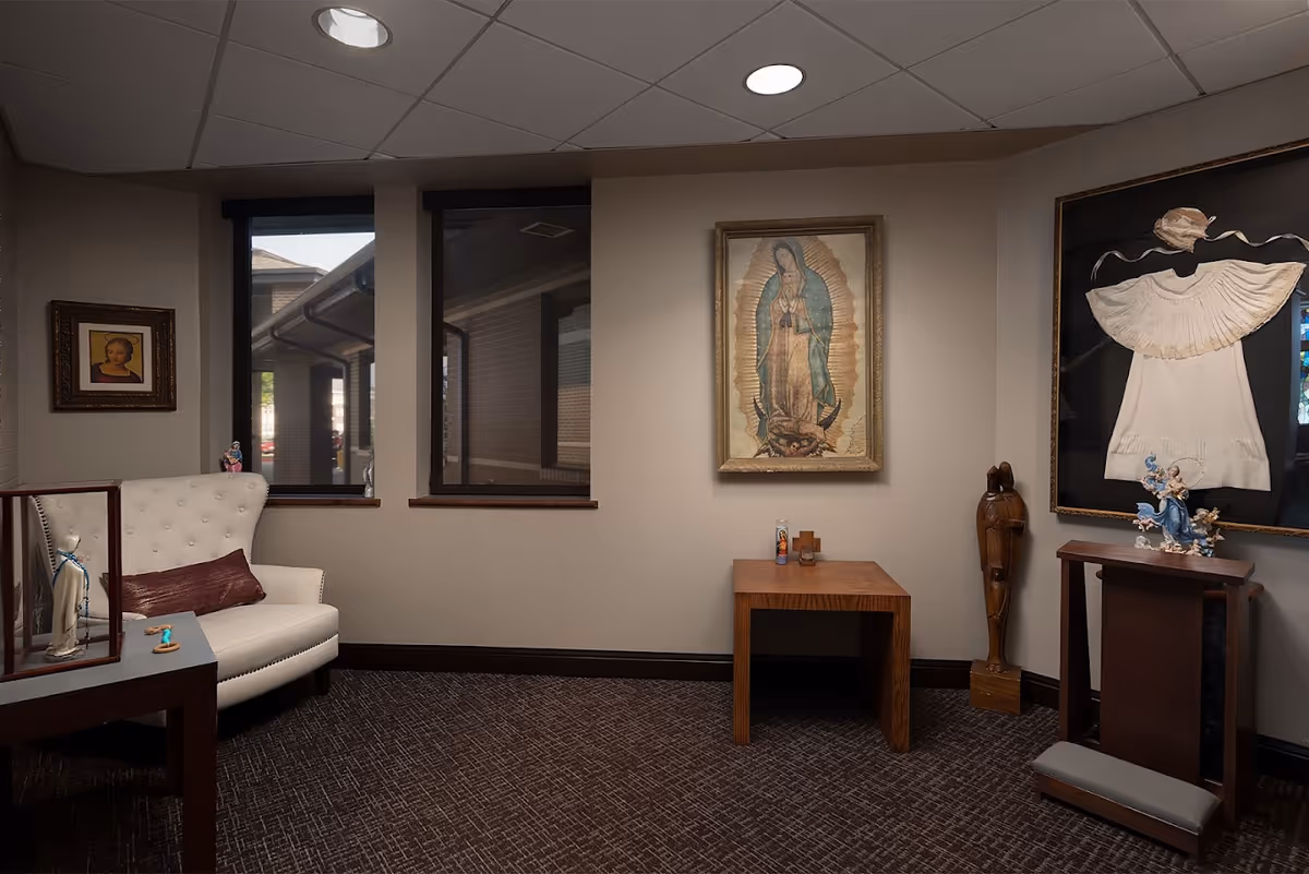 Interior quiet sitting room in a senior facility with a white upholstered chair, wooden tables, and religious artwork on the walls.