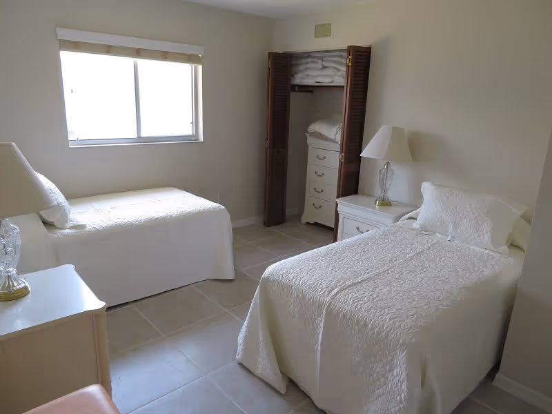 A simple bedroom with two single beds covered in white bedspreads. Each bed has a white pillow and a bedside table with a lamp. There is a window with blinds on the left wall and an open closet with shelves and folded linens on the back wall. The floor is tiled and the walls are painted light beige.