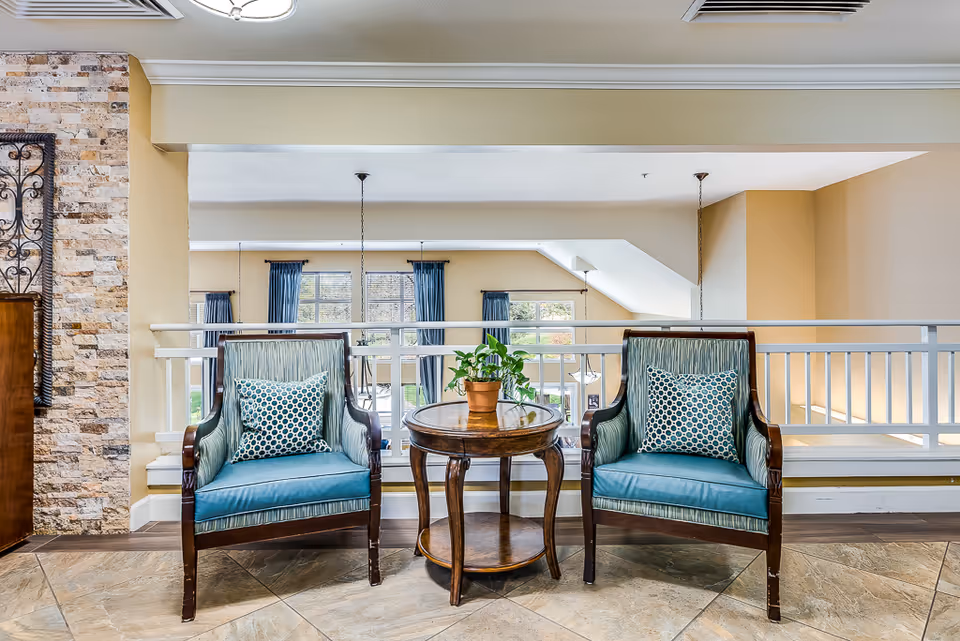 Two teal upholstered armchairs with patterned pillows flank a round wooden table holding a potted plant in a bright indoor seating area.