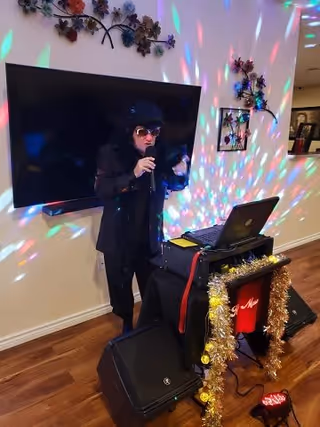 A person dressed in a black outfit and sunglasses is singing into a microphone in a room with colorful disco lights projected on the walls. There is a large flat-screen TV mounted on the wall behind them and a laptop on a decorated stand with gold tinsel and string lights. The room has wooden flooring and some wall decorations.