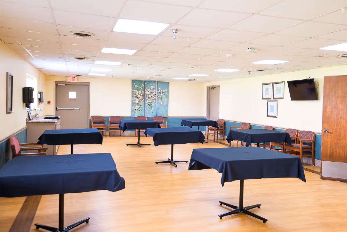 Empty multipurpose/dining room with several tables draped in dark tablecloths, chairs along the walls, wall art and a mounted TV.