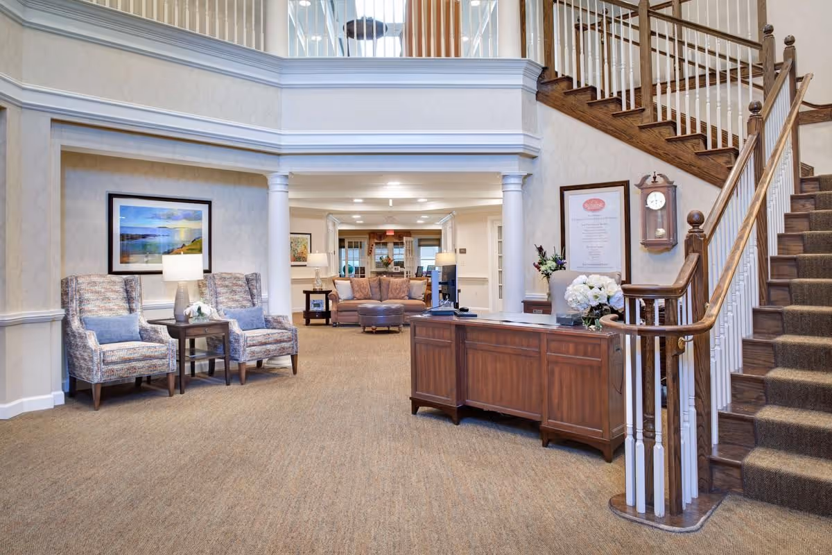 Interior view of a senior living facility lobby area with a wooden reception desk adorned with a flower arrangement, two patterned armchairs with a side table and lamp, a staircase with wooden handrails and carpeted steps, and a seating area with a sofa and ottoman in the background.