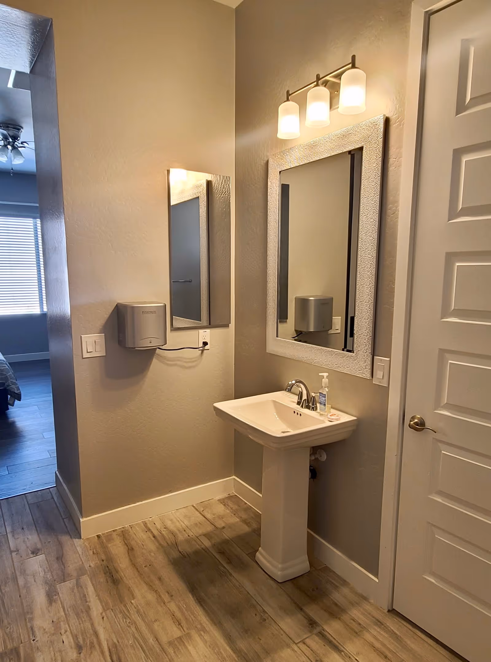 A small bathroom area with a white pedestal sink, a rectangular mirror with a textured white frame above it, and a three-light fixture mounted on the wall. To the left of the sink is a smaller mirror and a wall-mounted paper towel dispenser. The floor has wood-like tiles, and a white door with a silver handle is visible on the right. In the background, part of a bedroom with a window and blinds can be seen.