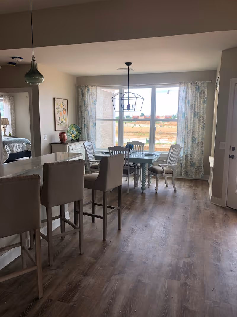 Interior view of a dining area with a rectangular table surrounded by six chairs near a large window with floral curtains. To the left, there is a kitchen counter with three bar stools. A bedroom is partially visible through an open doorway on the left side. The floor is wooden, and a modern pendant light hangs above the dining table.