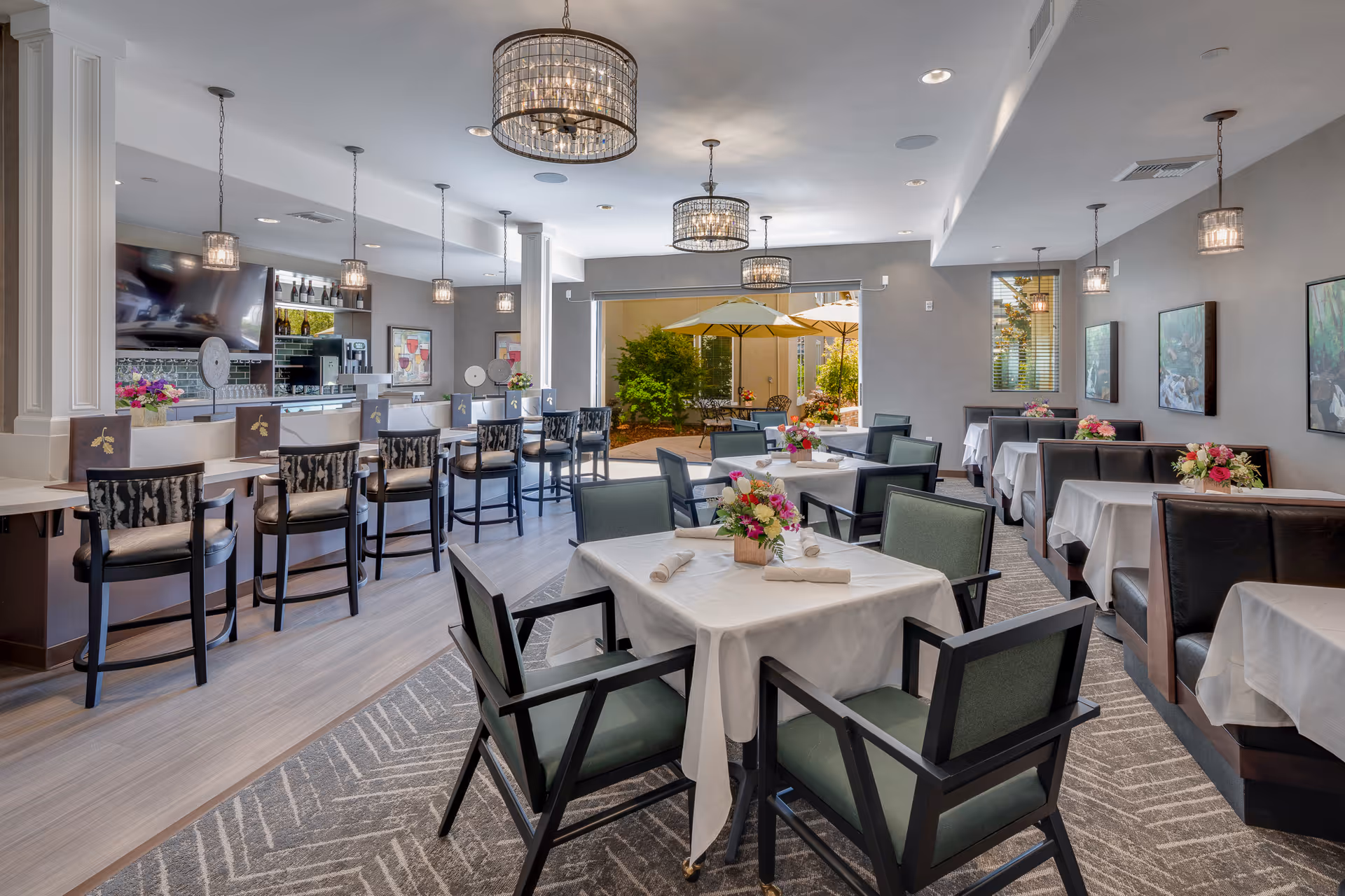 Bright dining room with tables set in white linens, upholstered chairs, a bar with stools, hanging chandeliers, and an open patio view.