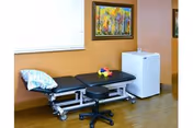 A therapy or examination room with a black adjustable medical table, a black rolling stool, a small white refrigerator, and a colorful painting on the orange wall. There is a window with closed blinds above the medical table.