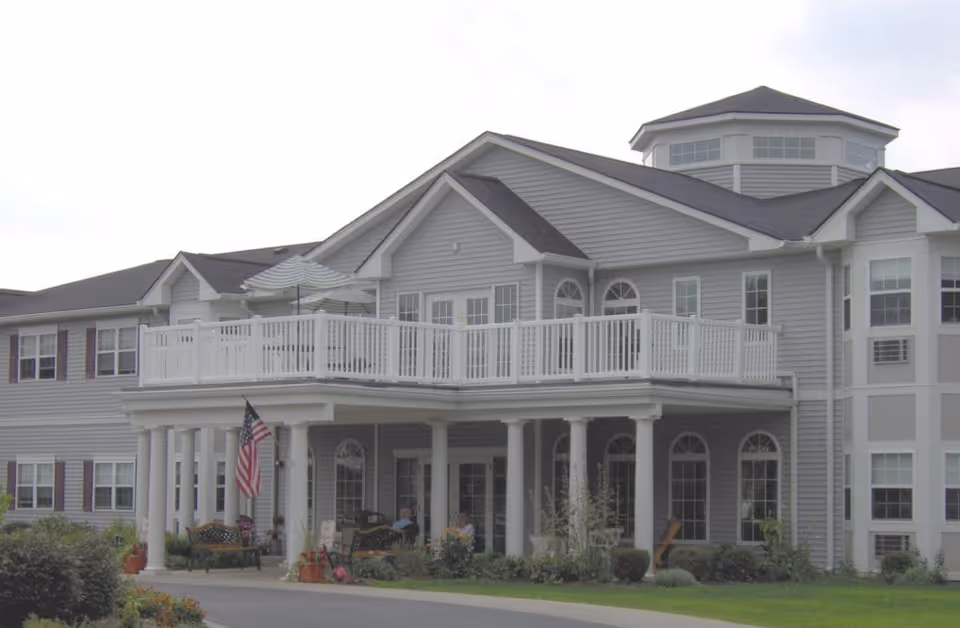 Exterior front of a two-story gray senior living building with white columns, a second-floor balcony, and an American flag.