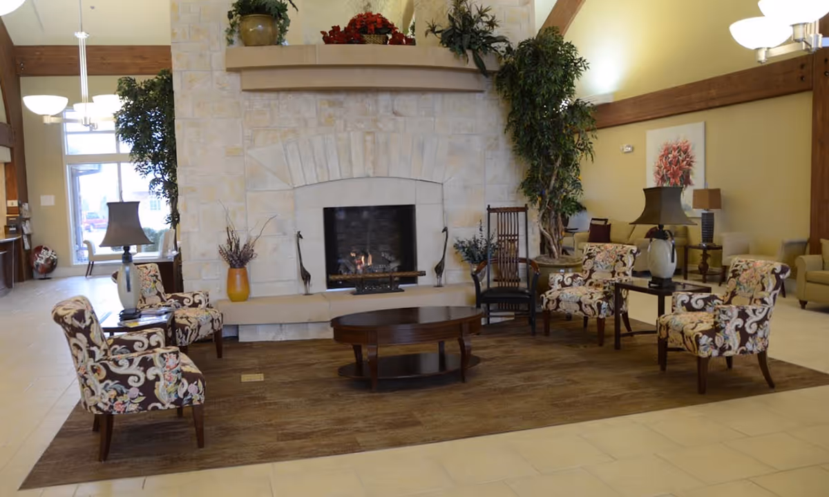 A cozy living room area in Silvercrest at Woodlands Creek featuring a stone fireplace with a fire burning, surrounded by four floral-patterned armchairs and a wooden coffee table on a brown rug. The room has beige tiled floors, potted plants, table lamps, and warm yellow walls with wooden beams.