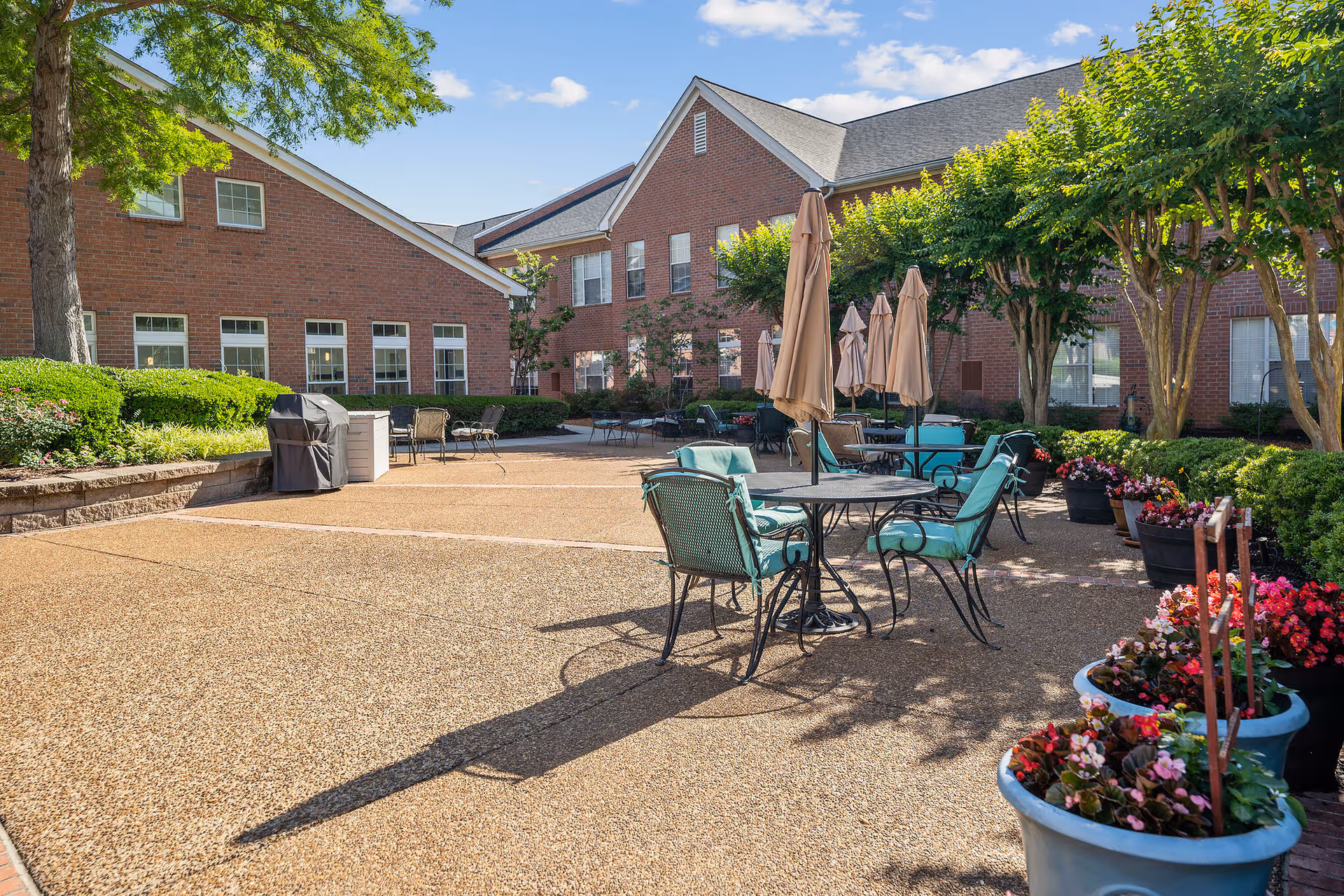 Outdoor patio area at Town Village Audubon Park with several tables and chairs featuring teal cushions, closed beige umbrellas, potted flowers, and trees. The background shows a brick building under a blue sky with some clouds.