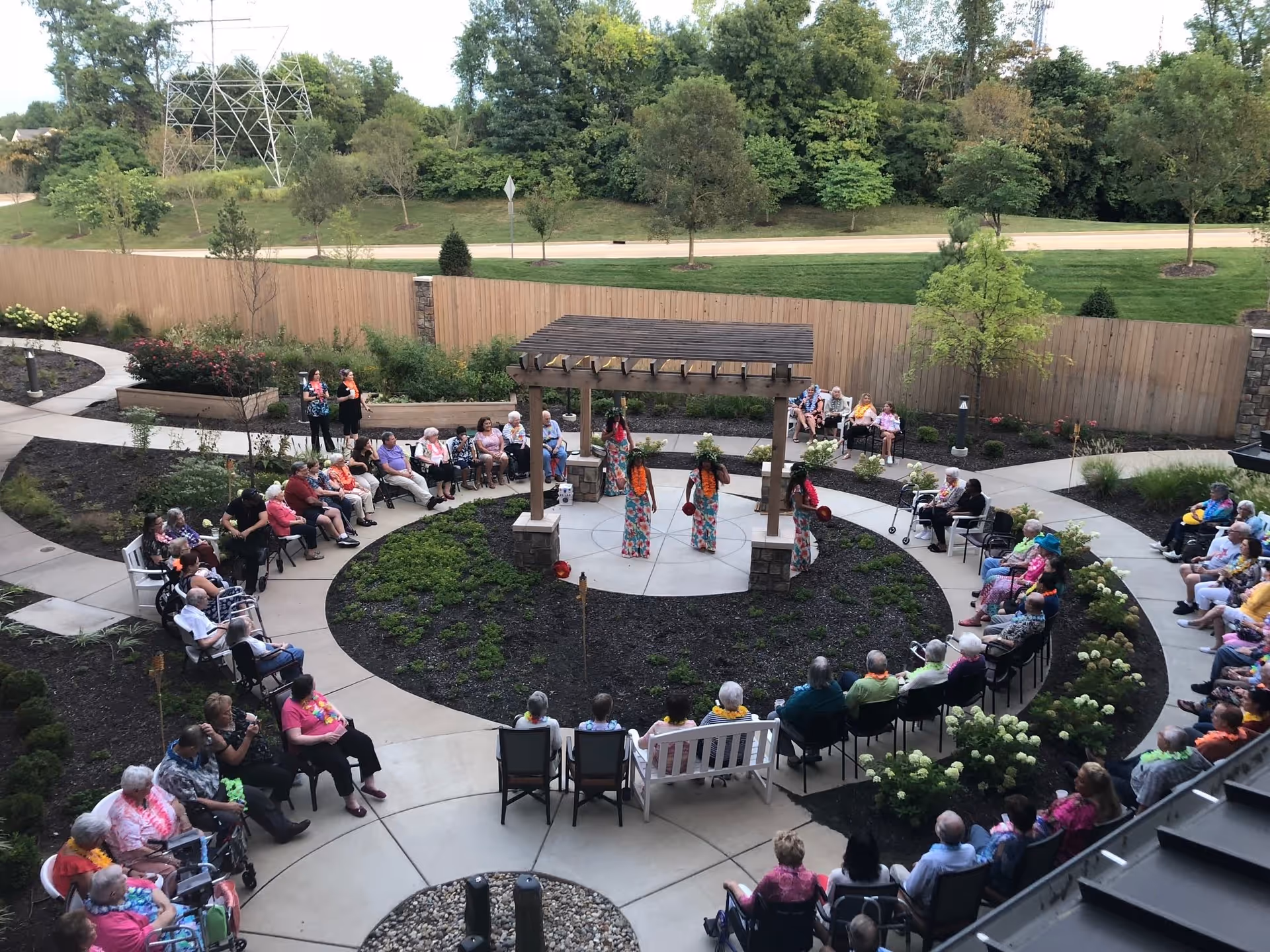 An outdoor courtyard at a senior living facility with a circular seating arrangement around a central pergola where three performers dressed in colorful outfits are entertaining a large group of elderly residents seated on chairs and benches. The area is landscaped with plants and trees, and a wooden fence and greenery are visible in the background.
