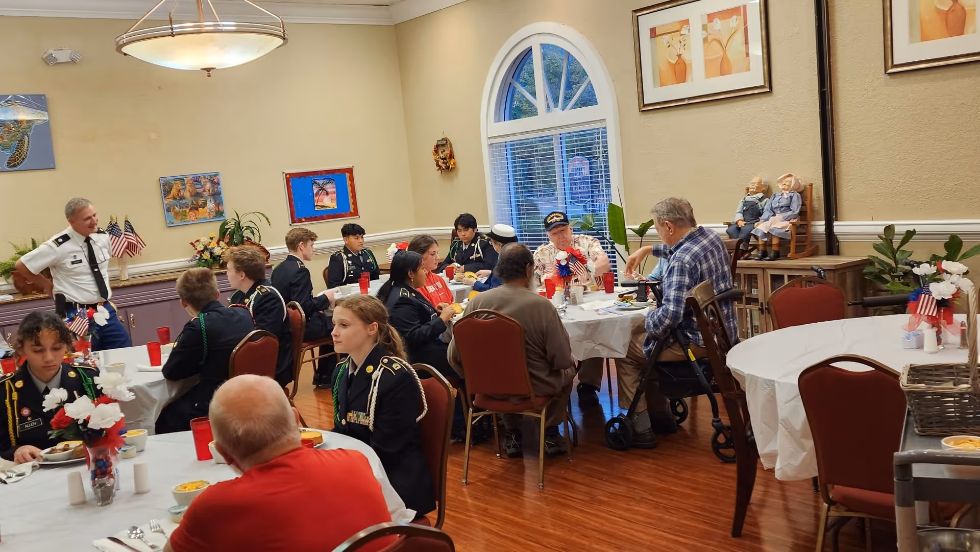 A group of people, including several young individuals in uniform and elderly individuals, are seated around tables in a dining room decorated with patriotic-themed centerpieces and artwork on the walls. The room has wooden floors, arched windows, and a warm, inviting atmosphere.