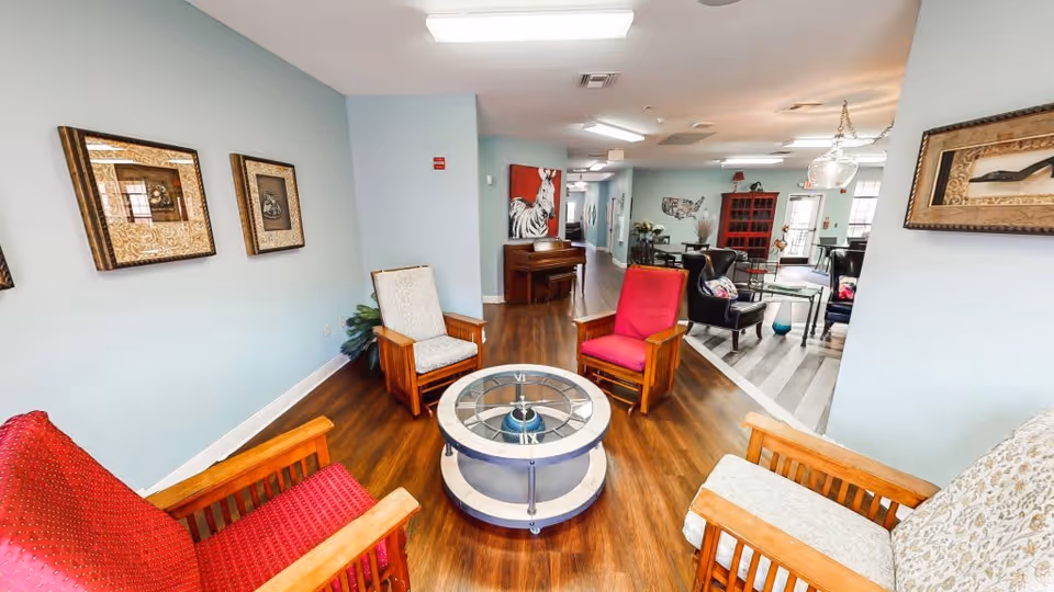 A cozy assisted living common area with wooden flooring and light blue walls. The room features four wooden armchairs with cushions arranged around a round glass-top coffee table. The chairs have different colored cushions, including red and beige. The walls are decorated with framed artwork. In the background, there is a piano against the wall and another seating area with black chairs and a red cabinet. The space is well-lit with ceiling lights.