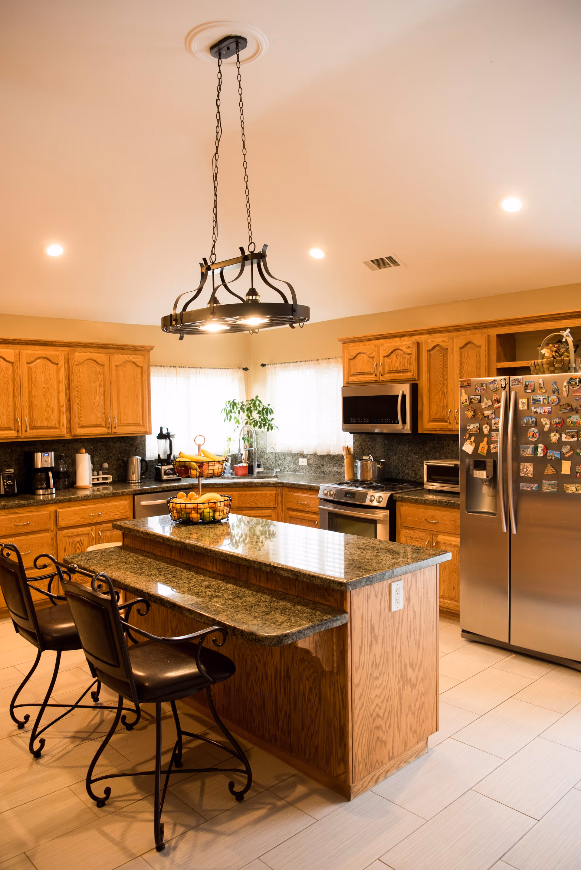 A spacious kitchen with wooden cabinets, granite countertops, and a central island with two wrought iron chairs. The kitchen features stainless steel appliances including a refrigerator covered with magnets, a microwave, and a stove. There is a hanging light fixture above the island and windows with sheer curtains letting in natural light.