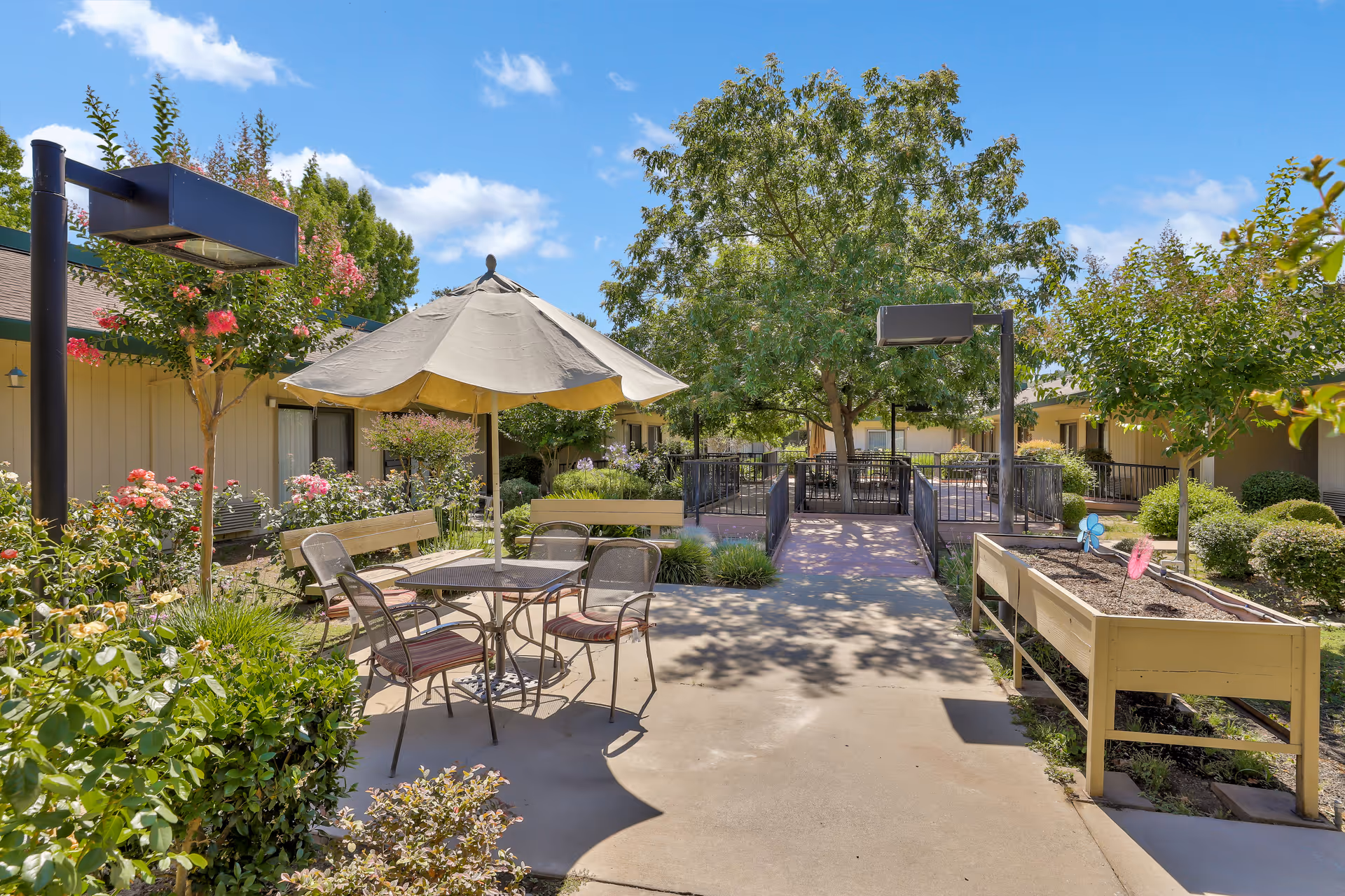 Outdoor courtyard area at Park Merced Assisted Living with a round table and four chairs under a large beige umbrella. There are benches, flowering plants, trees, and raised garden beds along a paved walkway. The sky is clear with a few clouds.