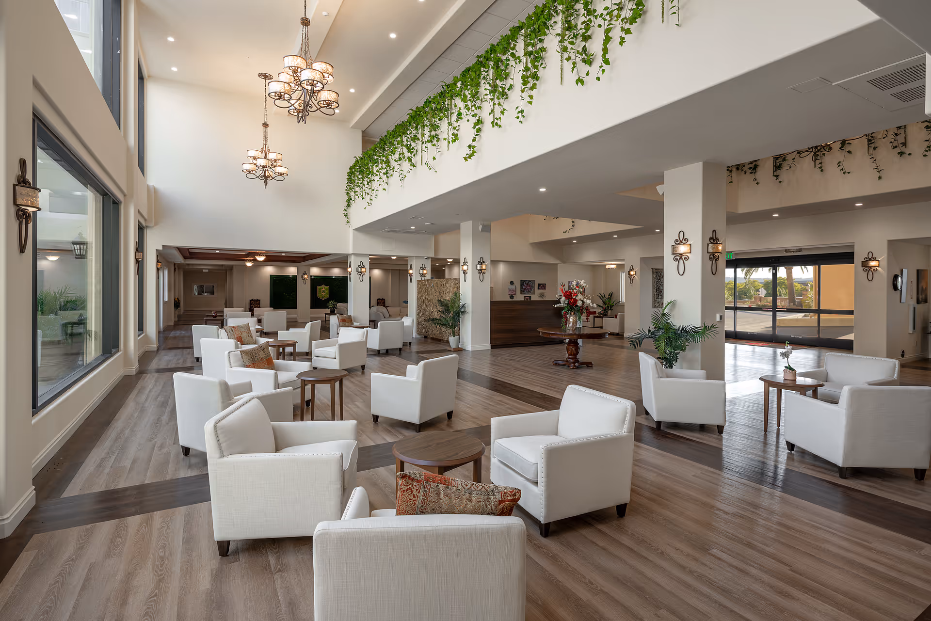 Spacious senior living facility common area with multiple white armchairs arranged around small wooden tables on a wood floor. The room features high ceilings with hanging chandeliers, large windows, decorative plants hanging from the upper level, and a reception desk in the background.