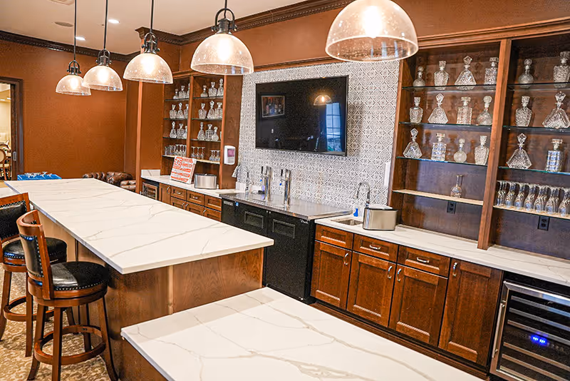A communal kitchen/bar area with marble-topped islands, pendant lights, wooden cabinets and glassware on display.