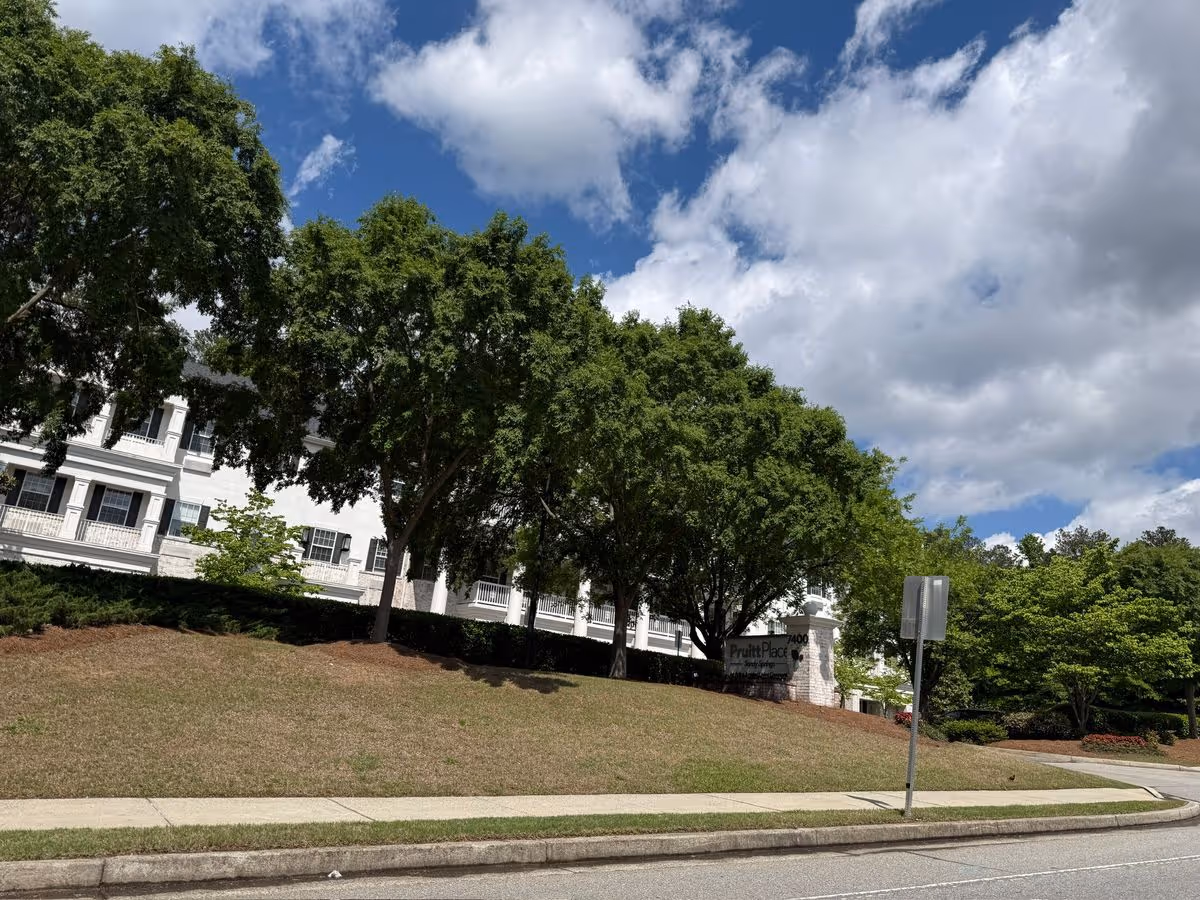 Front exterior of a white multi-story senior living building partially hidden by large trees with a lawn and a PruittPlace sign under a blue sky.