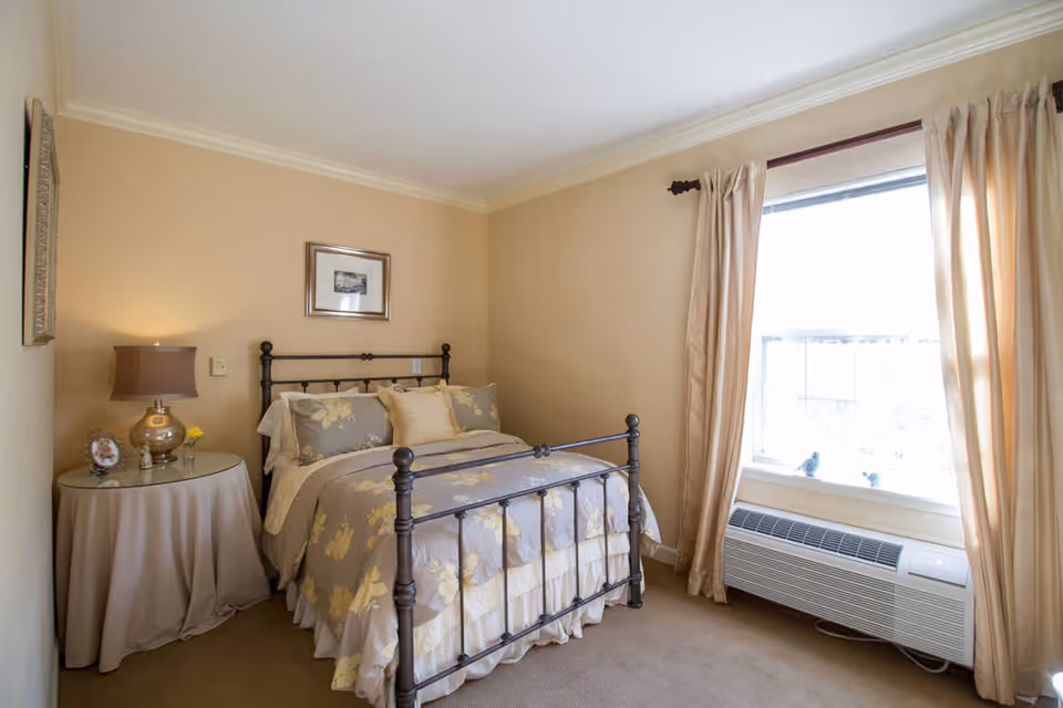 A cozy bedroom with a metal frame bed dressed in floral-patterned bedding in shades of gray and yellow. There is a round side table covered with a beige cloth, holding a lamp, a small framed photo, and a small vase with a yellow flower. The room has beige walls, a window with beige curtains, and a white air conditioning unit below the window.