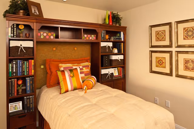 A cozy bedroom featuring a single bed with a beige comforter and multiple decorative pillows in orange, yellow, and striped patterns. The bed is framed by a wooden headboard with built-in shelves filled with books, baskets, and decorative items. On the wall to the right, there are four framed square artworks with intricate designs. The room has a warm and inviting atmosphere.