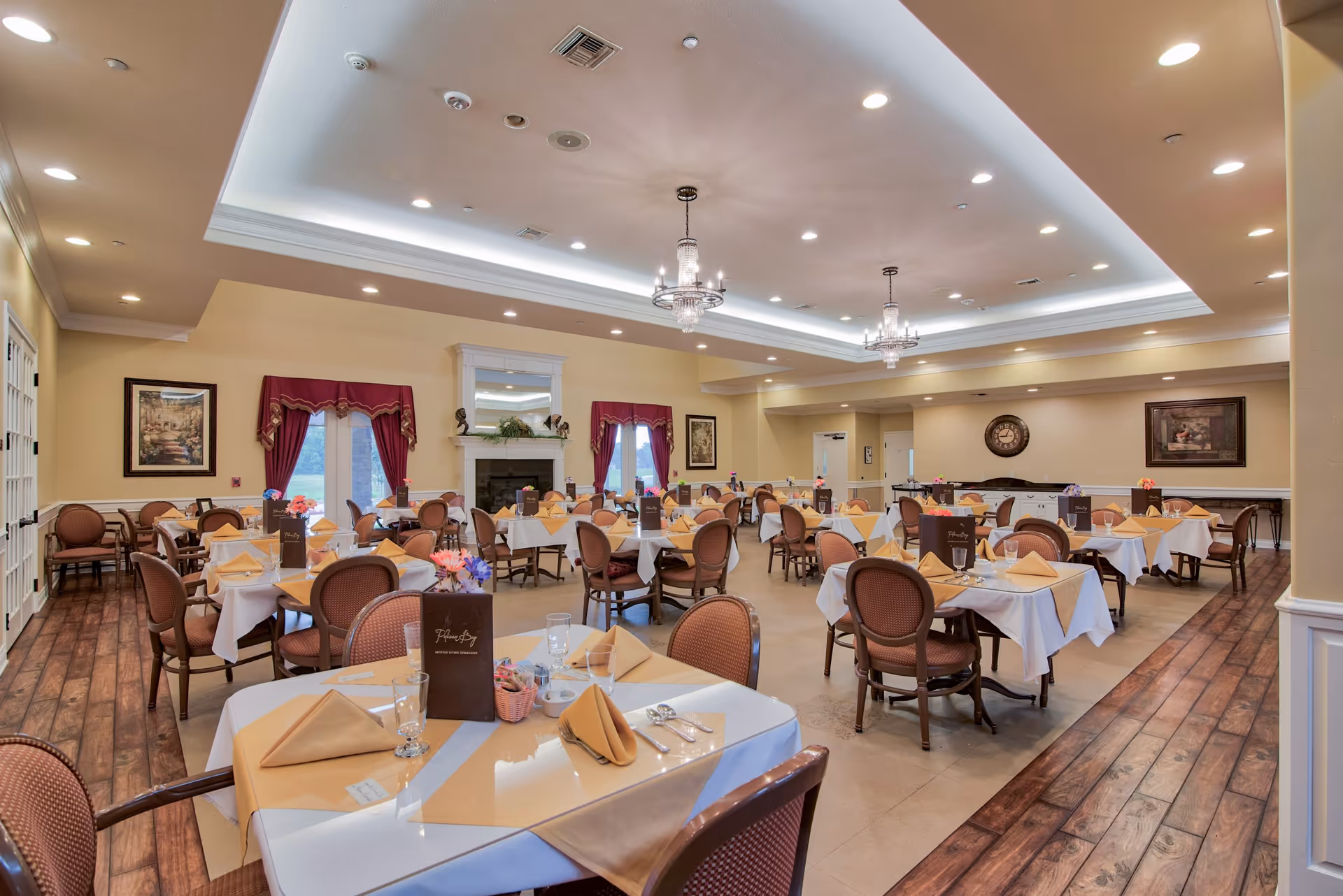 A spacious dining room in Pelican Bay Assisted Living Community with multiple round tables covered with white tablecloths and beige napkins. Each table is set with glasses, silverware, and a menu. The room features wooden flooring along the edges, a fireplace with a mirror above it, red curtains on the windows, framed artwork on the walls, and elegant chandeliers hanging from the ceiling.