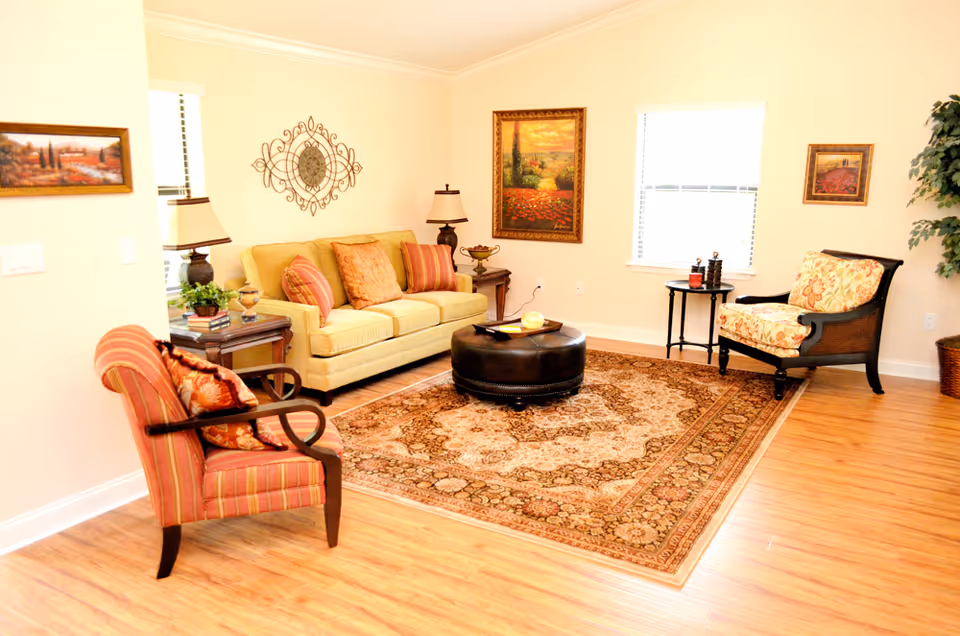 A bright living room with a beige sofa adorned with multiple cushions, two armchairs with patterned upholstery, a round dark brown ottoman on a large ornate area rug, wooden flooring, and several framed paintings on the cream-colored walls. There are two windows with blinds and a potted plant in the corner.