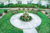 A circular outdoor garden area with a three-tier stone fountain in the center, surrounded by a concrete walkway and green grass. There are three wooden benches placed around the fountain, and shrubs and trees line the background near a white building.