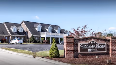 Exterior view of Chandler House Assisted Living Community building with a brick sign in the foreground displaying the community name and address. The building has a pitched roof with dormer windows and a parking area with cars visible.