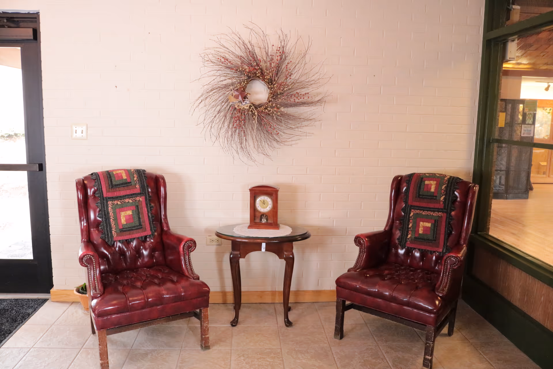 Two red leather armchairs with patterned throws draped over the backs, positioned on either side of a small wooden table with a clock on it, against a white brick wall decorated with a wreath made of twigs and berries.