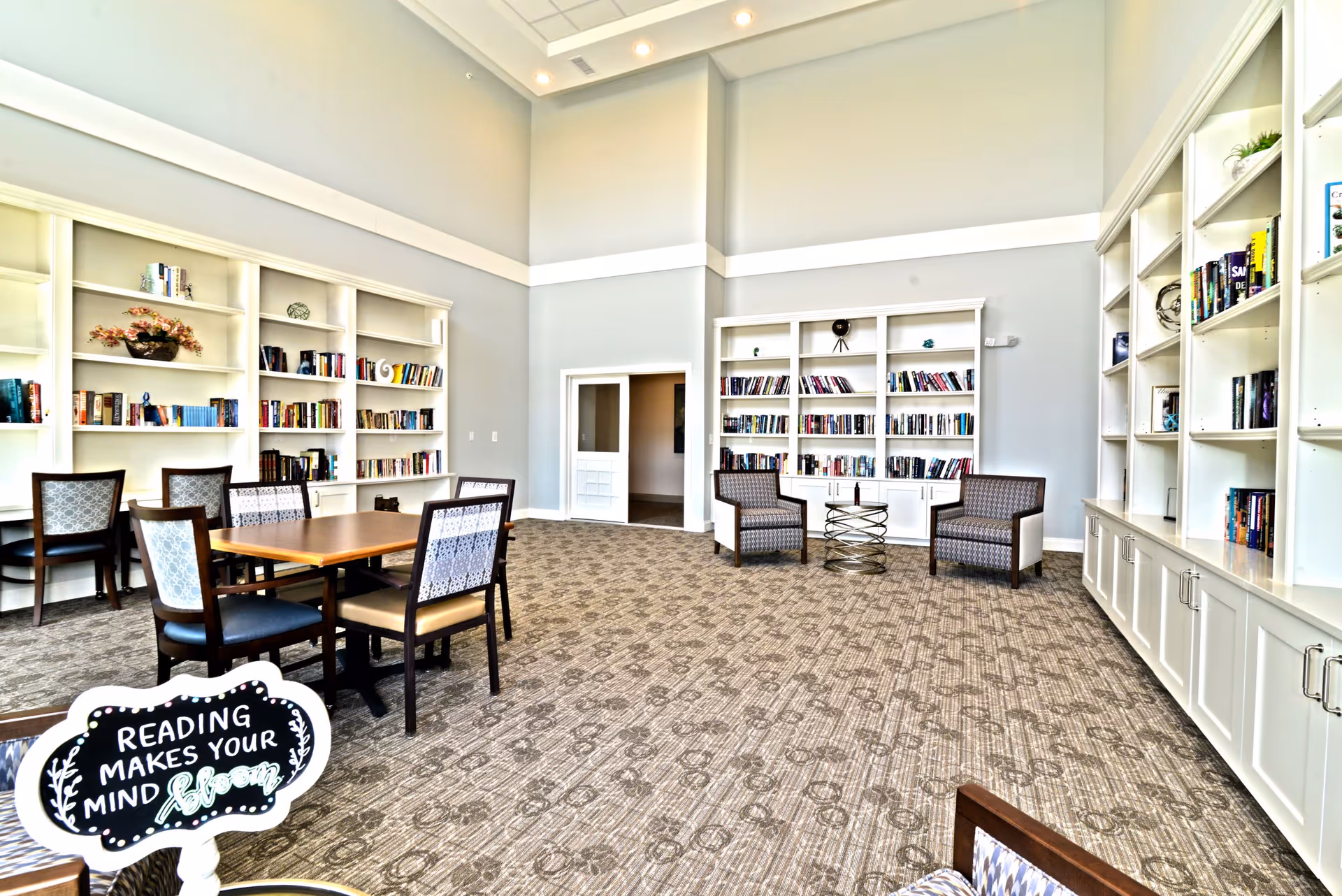 A spacious and well-lit reading room with tall ceilings, carpeted floor, and light gray walls. The room features multiple white bookshelves filled with books and decorative items. There are several chairs and tables arranged for reading or socializing. A small sign in the foreground reads 'Reading makes your mind bloom.'