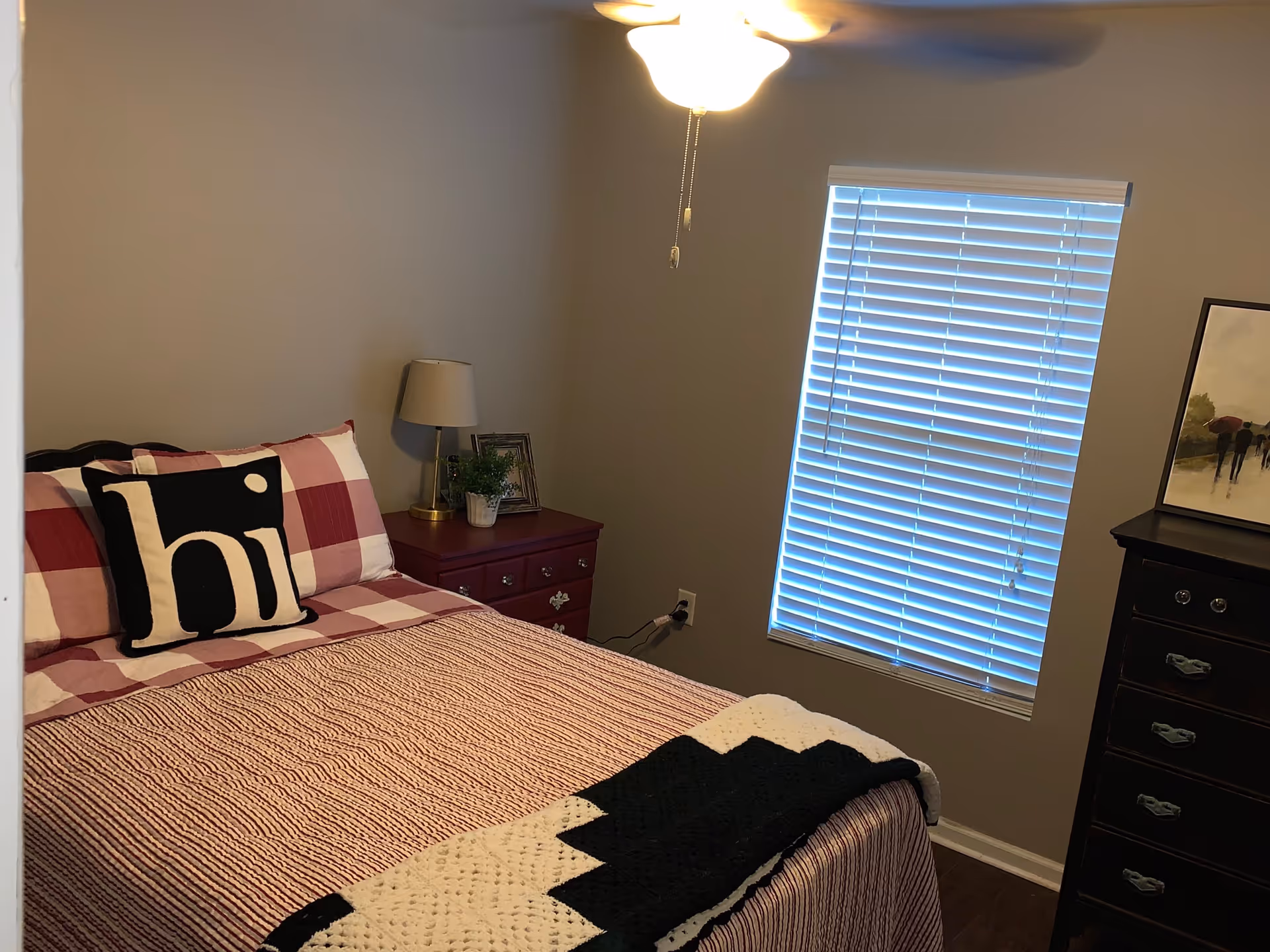 A cozy bedroom with a bed covered in red and white checkered bedding and a black and white pillow with the word 'hi'. There is a red nightstand with a lamp, a small plant, and a framed picture. A window with closed blinds is on the right wall, and a dark wooden dresser with a framed painting on top is next to the window. A ceiling fan with a light is on the ceiling.