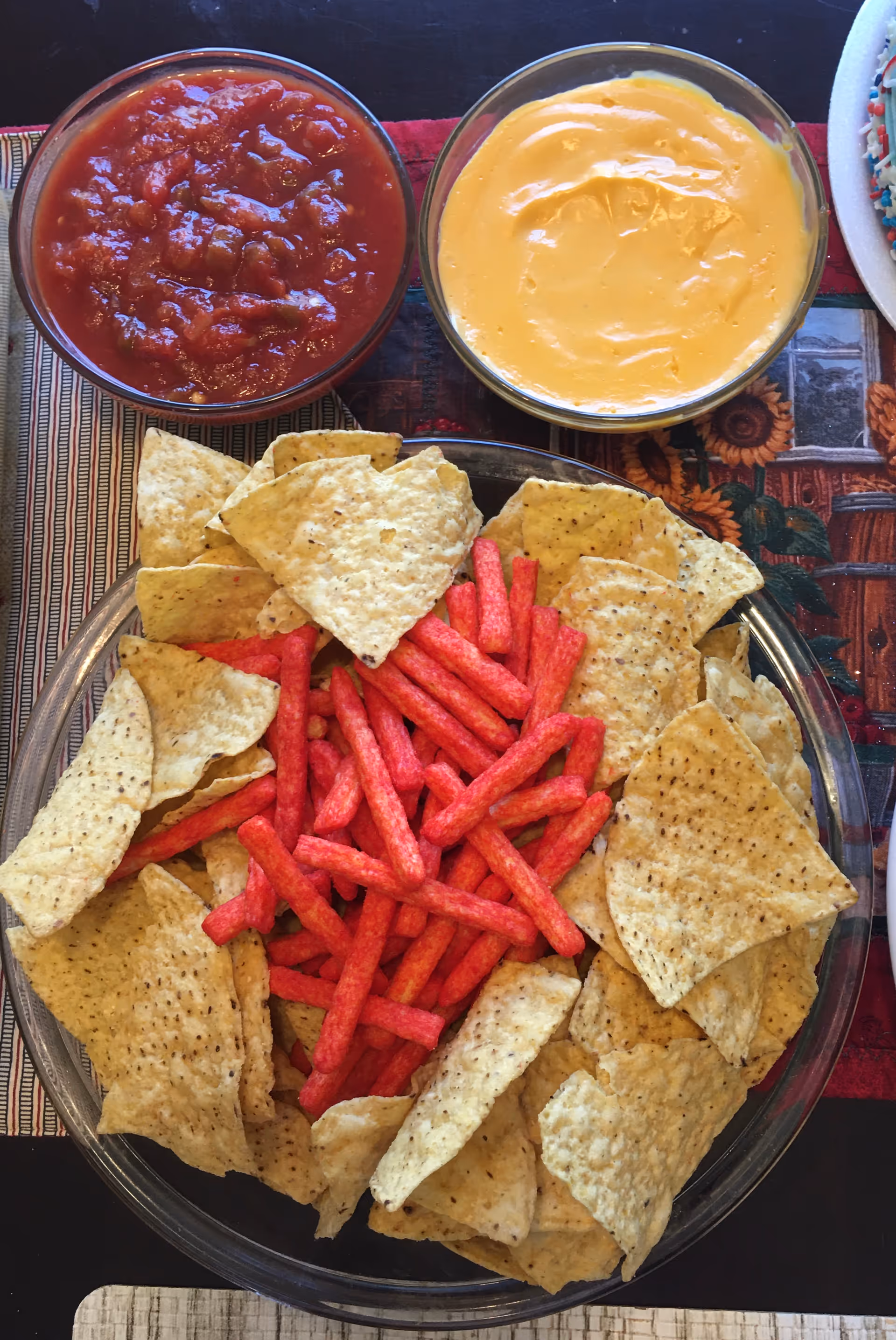 A clear glass bowl filled with tortilla chips and red spicy snack sticks, accompanied by two small bowls of dips, one with salsa and the other with cheese sauce, placed on a patterned tablecloth.
