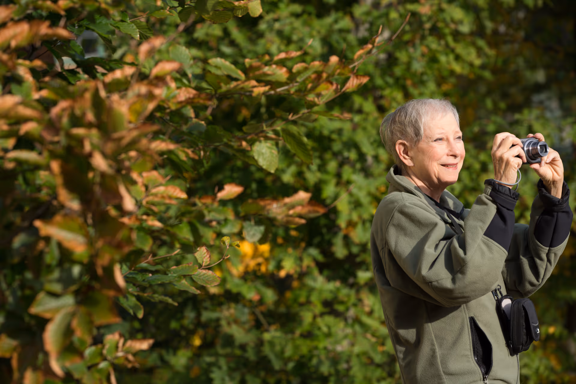 An elderly woman wearing a green jacket is outdoors taking a photo with a small camera, surrounded by green foliage and trees.