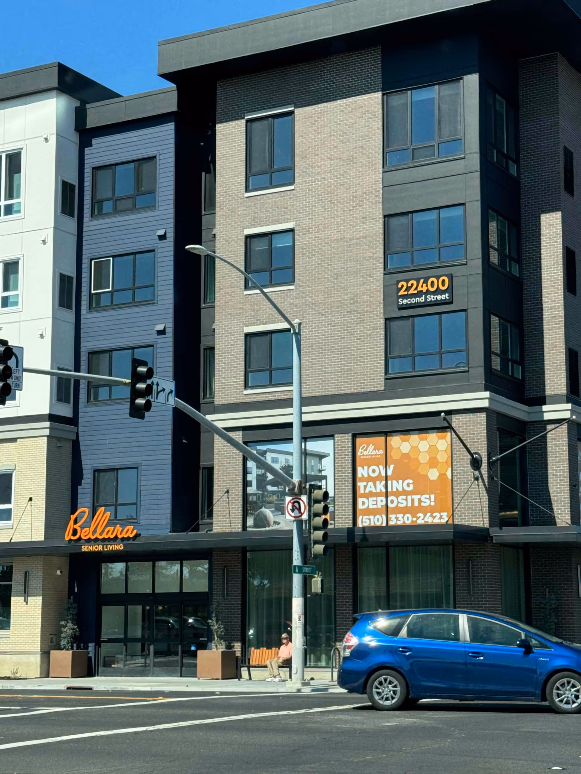 Exterior view of Bellara Senior Living building at a street corner with a traffic light and a blue car parked in front. The building has multiple floors with large windows and signs displaying the address 22400 Second Street and a banner stating 'Now Taking Deposits!' with a phone number. A person is sitting on a bench near the entrance.