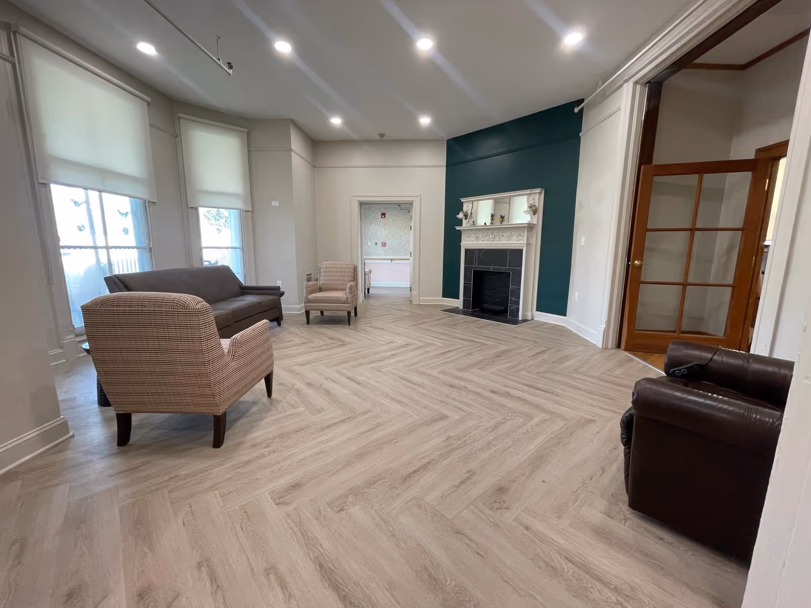 A spacious living room with light wood herringbone flooring, a dark green accent wall with a white fireplace and mirror above it, a brown leather armchair, a plaid armchair, and a dark gray sofa. The room has large windows with white roller shades and recessed ceiling lights. A wooden door with glass panels is visible on the right side.
