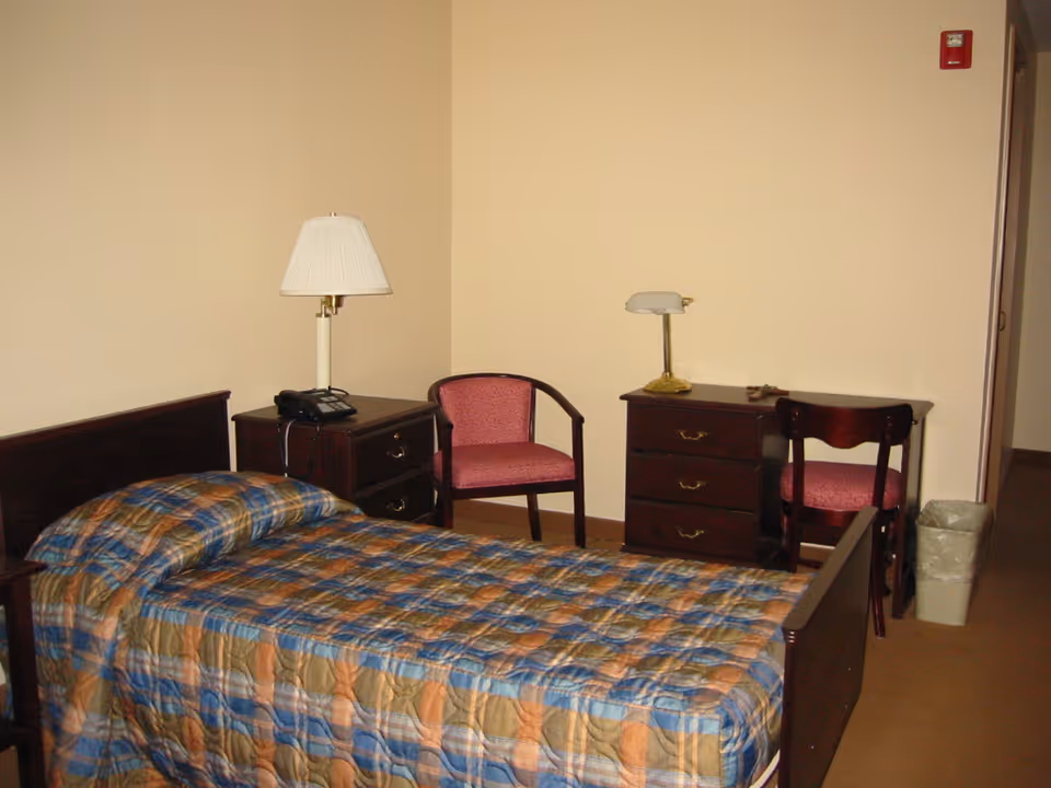 Furnished single-occupancy bedroom with a bed, nightstand, desk and two chairs against a beige wall.