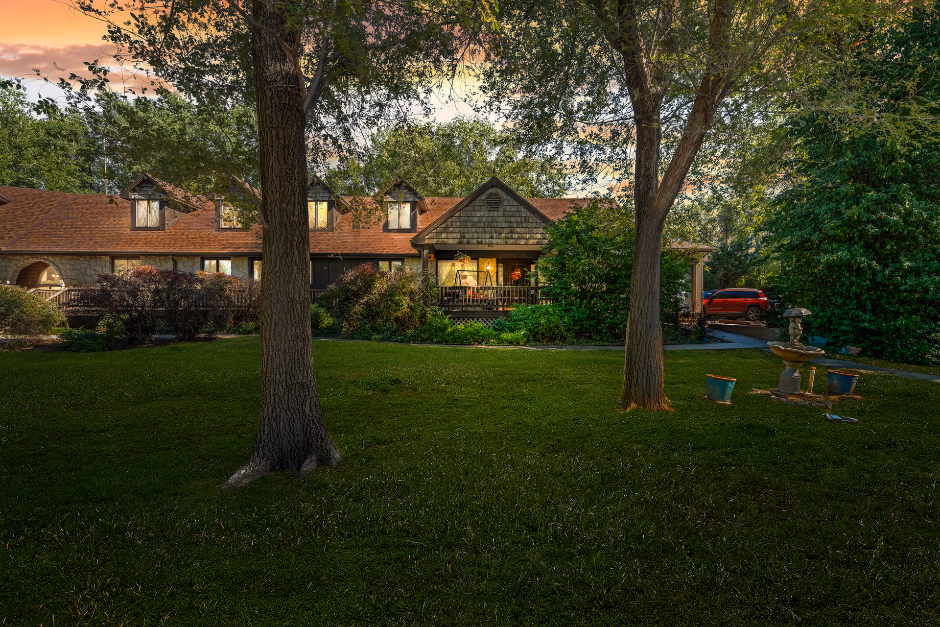 Front exterior of a cottage-style building at dusk with a lawn, trees, a lit porch, and a fountain.