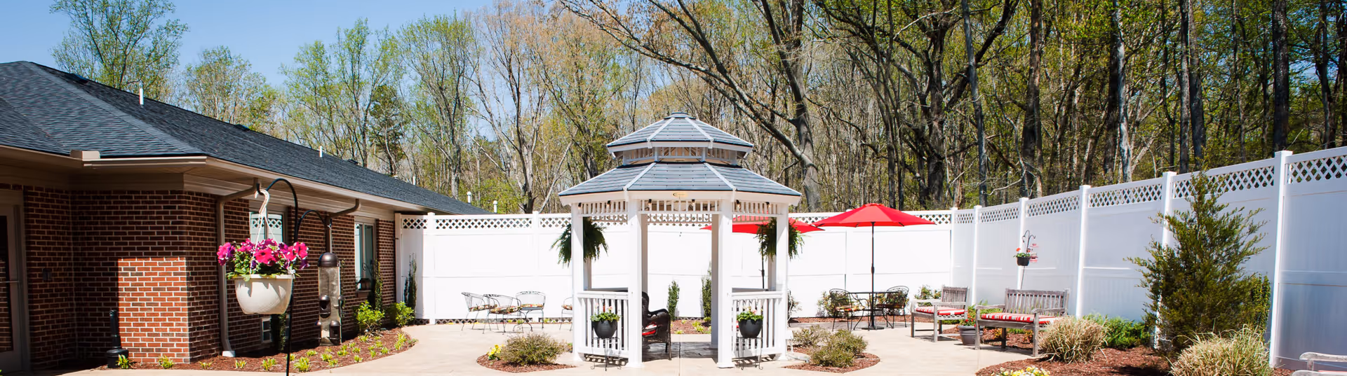 Outdoor courtyard area at Commonwealth Senior Living At South Boston featuring a white gazebo in the center, surrounded by benches, tables with red umbrellas, hanging flower pots, and a white privacy fence with trees in the background.