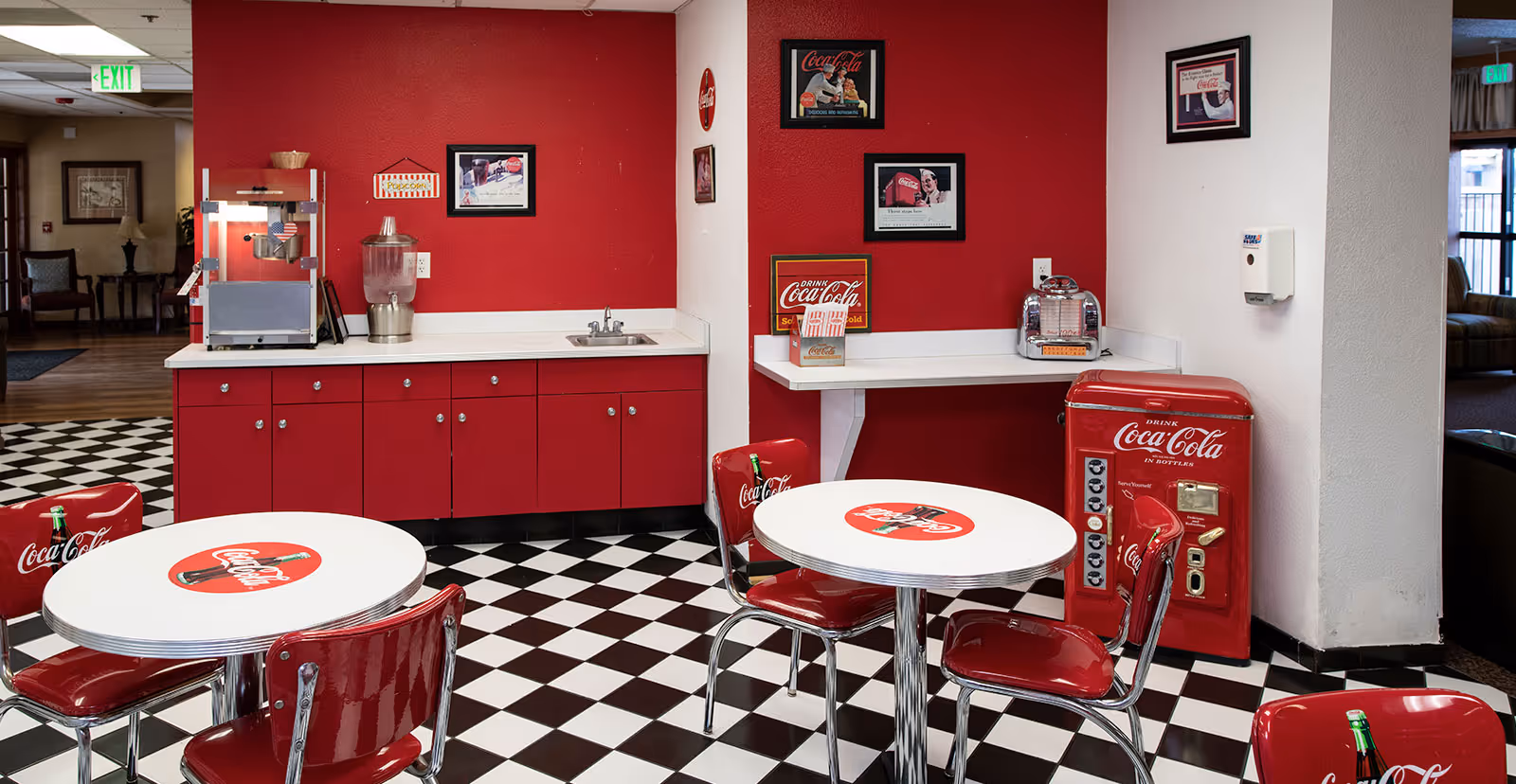 Retro Coca-Cola themed dining area with red cabinets, round tables and chrome chairs on a black-and-white checkered floor.
